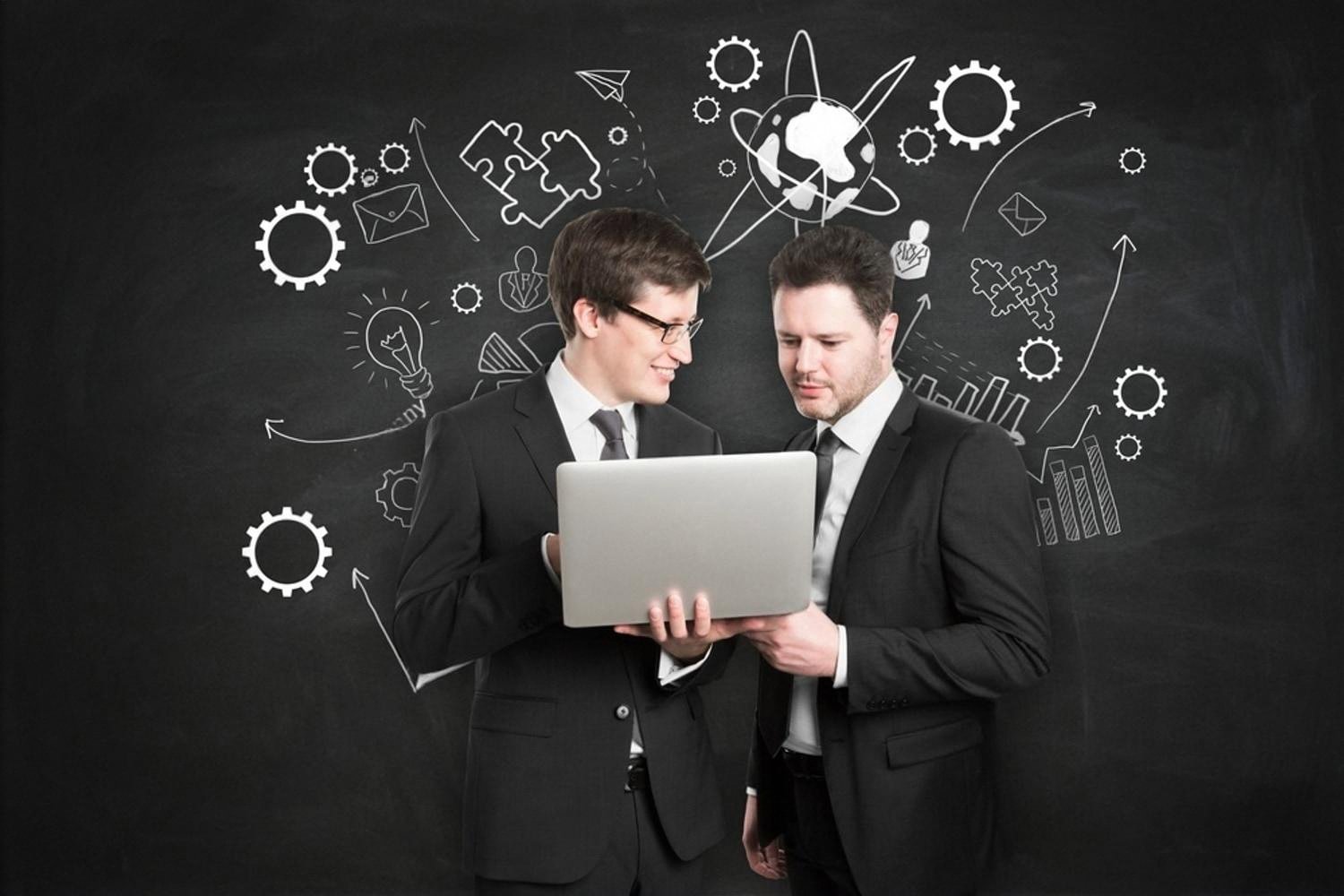 Two specialists reviewing a laptop in front of a chalkboard covered with knowledge and skills icons